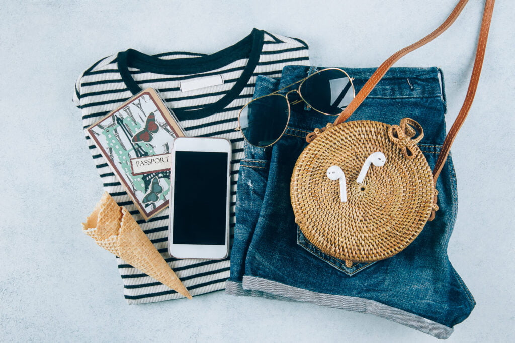 Summer women's clothing and accessories flat lay: denim shorts, striped shirt, sunglasses, and straw bag