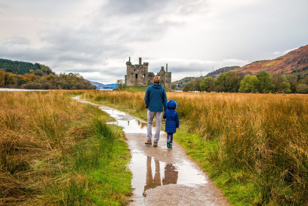 a person and a child walking on a dirt road with a castle in the background