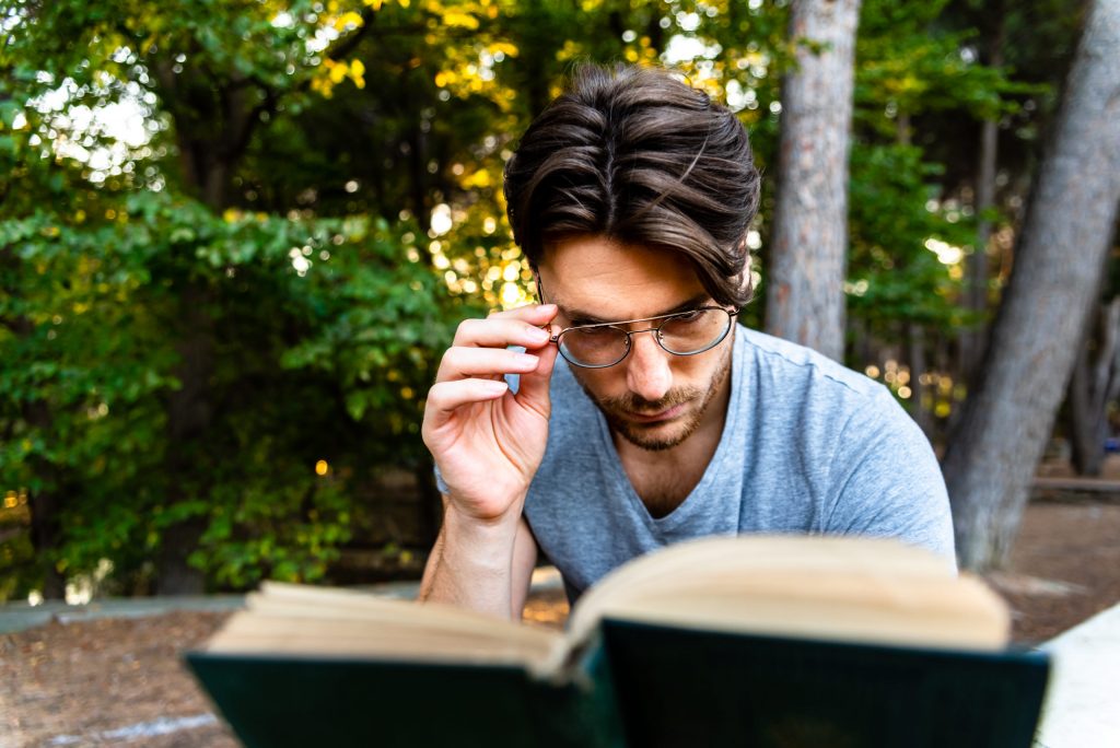 Male student with glasses carefully reads an old book in a park